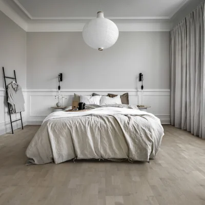 Minimalist bedroom with a large cream duvet bed centre, two nightstands and lamps, white paneled walls, pale wood floor, and grey curtains.