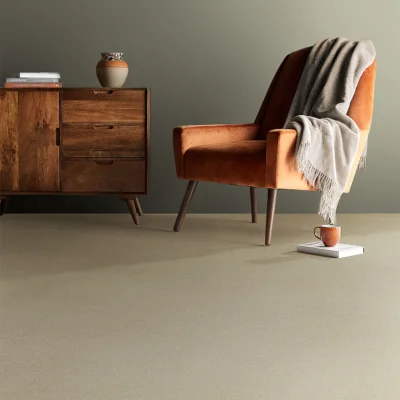 Warm-toned living corner featuring a burnt-orange armchair with a grey throw, a wooden dresser, a vase, and a mug on a book on a beige carpet.