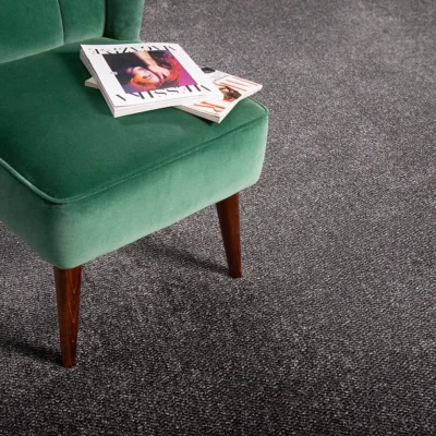 Green upholstered chair with wooden legs on a grey speckled carpet; magazines stacked on the seat.