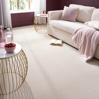 Bright living room with a cream sofa, pink throw and cushion, beige carpet and gold wire-frame side tables; a book on the floor and a bowl of berries on a table.
