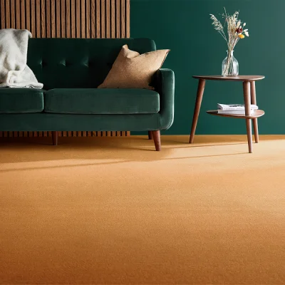 Emerald green velvet sofa with a cream throw and beige cushion, on an orange carpet, beside a round wooden side table with a vase of dried flowers.