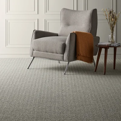 Grey armchair with metal legs on a carpet; a rust throw over the arm, beside a small wooden table with magazines and a vase of dried flowers.