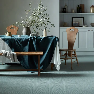 A dining space with a dark teal draped table, a wooden bench, and a vase of olive branches; a wooden chair and pale cabinetry in the background.