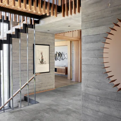 Interior with concrete walls, wooden ceiling slats, a black metal staircase, framed art, and a sunburst mirror on the right; doorway leads to a room.