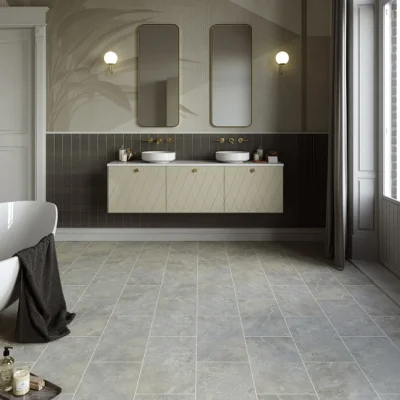 Modern bathroom with a floating beige double vanity, two vessel sinks and brass taps, dark tiled wall, two rounded mirrors, wall sconces, grey floor tiles, bathtub left, window right.