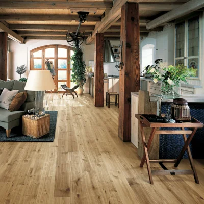 Bright, rustic open-plan living area with exposed timber beams and light wooden floors; grey sofa with cushions, floor lamp, potted plants, arched window at the back, and a rear kitchen.