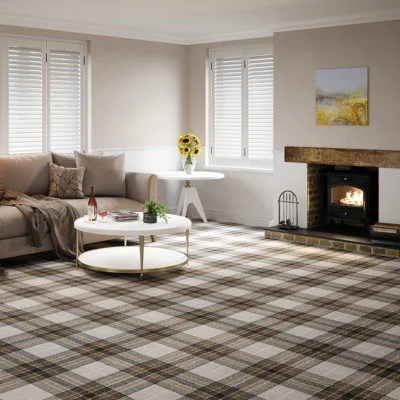 Bright living room with beige walls and white shutters; plaid carpet, beige sofa, round coffee table with wine and magazines, a vase of sunflowers, and a lit fireplace with a painting above.