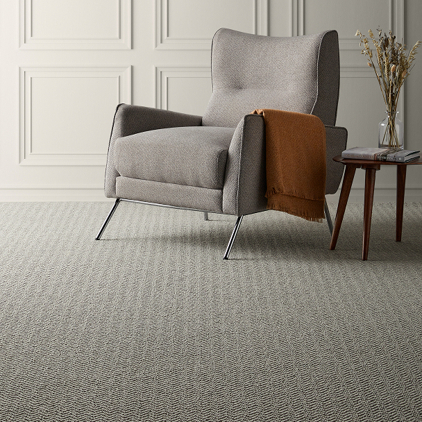 Grey upholstered armchair with slim metal legs in a neutral paneled room. A brown throw over the arm, beside a wooden side table with a glass vase of dried stems.