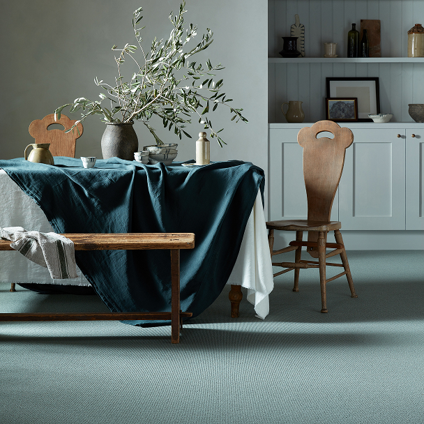 Rustic dining scene with a table draped in dark teal cloth, an olive branch arrangement in a ceramic vase, a wooden bench and chairs, and light cabinets in the background.
