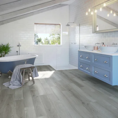 Bright modern bathroom with light wood flooring, blue freestanding tub, blue double vanity, white subway tiles, a window, plant and a towel on a stool.