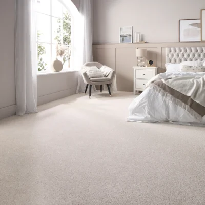 Bright neutral bedroom with a tufted headboard bed, white bedding, beige walls, a cushioned chair by a large arched window, and a white bedside table.