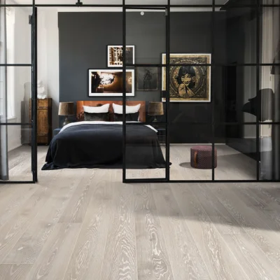 Bedroom seen through a black metal-framed glass partition; dark duvet bed with white/brown pillows, two lamps, and framed art on a charcoal wall.