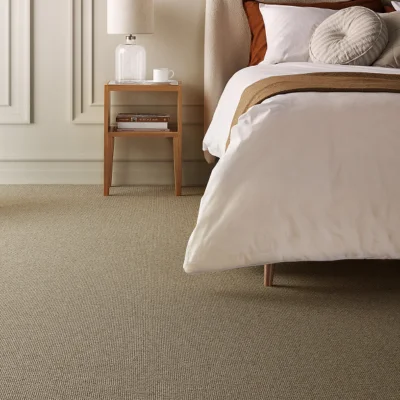 A neatly made bed with white linens and a beige throw, beside a wooden bedside table with a lamp, a cup and books on a beige carpet.