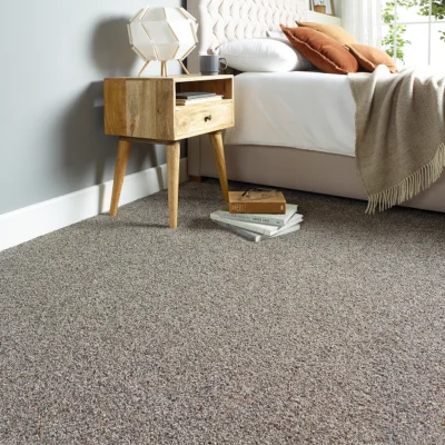 Cozy bedroom corner with a wooden nightstand and geometric lamp beside a white bed, rust cushions, a fringed throw, and a stack of books on the carpet.