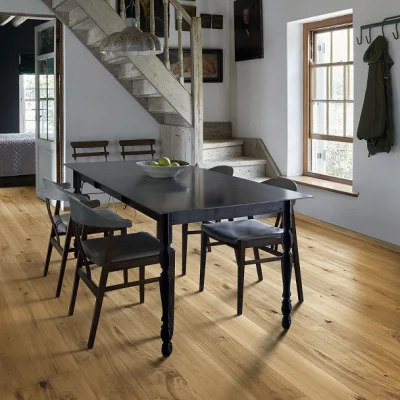 Rustic dining area with a dark wooden table and chairs, a bowl of apples, light wooden floor, a concrete staircase, and a large window with a coat rack nearby.