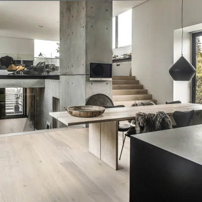 Open-plan modern interior with light wooden floors, concrete pillar, long dining table, fur throw, black pendant lamp, stairs and large windows.