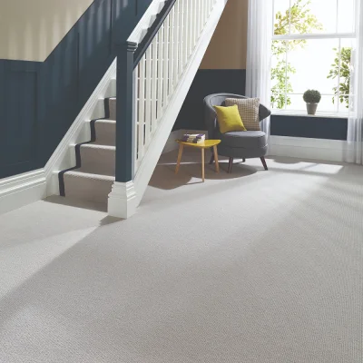 Bright living area with beige carpet, navy stair rail, grey armchair with yellow and check cushions, a yellow side table, and a window with white curtains and a potted plant.