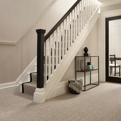 Neutral hallway with a white staircase and black handrail; a slim black metal console table holds decor, and a cushion rests on the carpeted floor.