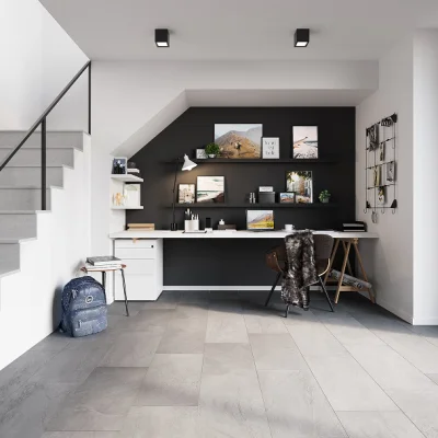 Home office under a staircase with a black wall, white desk, and shelves filled with photos and plants; chair with fur coat, backpack nearby.