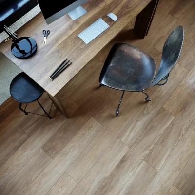Top-down view of a wooden desk with monitor, keyboard, mouse, scissors and pens; a worn leather chair and a small black stool on a wooden floor.