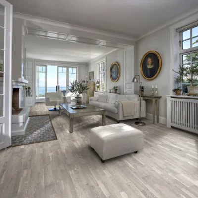 Bright coastal living room with pale walls and white sofas, a glass coffee table, and large windows framing a sea view; gold-framed portraits.