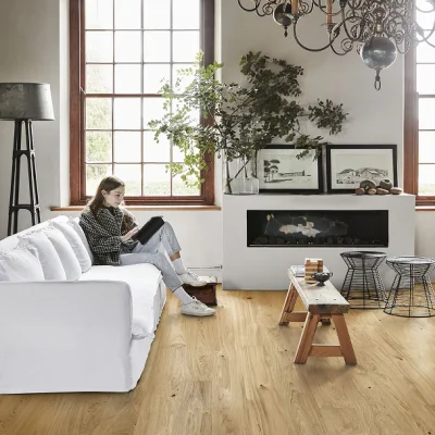 Person seated on a white sofa reading a book in a bright living room with a large window, a tall plant, a fireplace with framed photos above, and a wooden floor.