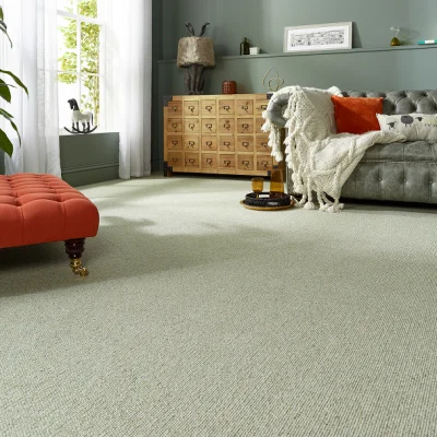 Living room with pale green walls, grey tufted sofa with orange cushion and cream throw, wooden chest of many drawers, coral ottoman, by a large window.