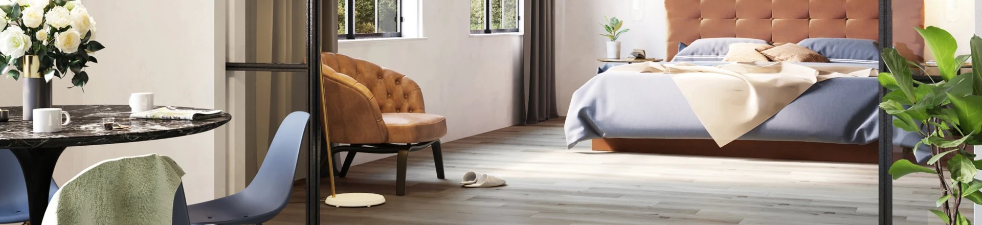 Open-plan room with a tufted headboard bedroom, a round marble dining table with white roses, blue chairs, a brown armchair, and green plants by a glass divider.