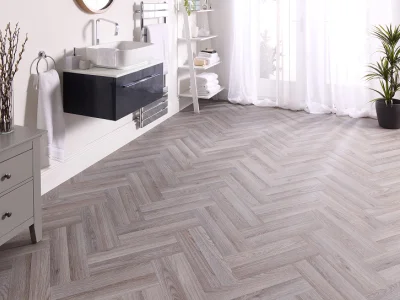 Bright, modern bathroom with pale herringbone wood floor, white sink and shelves, a black floating vanity, towels and a tall plant by sheer-curtained windows.