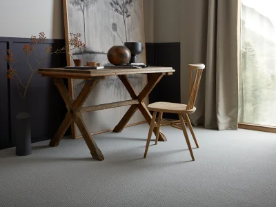 Rustic wooden desk and chair in a calm room, with a framed painting, pottery and dried branches; dark panelling, grey carpet, and a window.