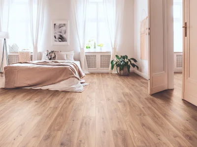 Bright, airy bedroom with a low beige bed, white sheer curtains, light wood flooring, a potted plant by the radiator, and an open door.
