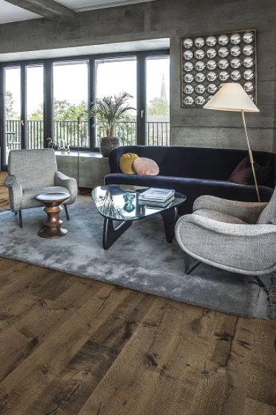 Modern living room with wood floors, windows and balcony; dark blue sofa, two chairs, glass coffee table on a rug, floor lamp and plants.