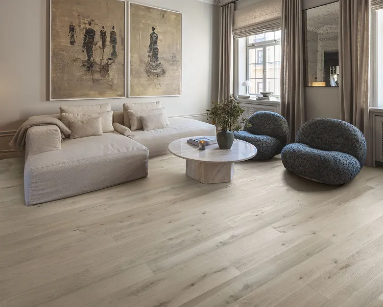 Bright living room with a pale beige sectional, two rounded blue chairs, round marble coffee table, plant, and large windows with beige curtains.