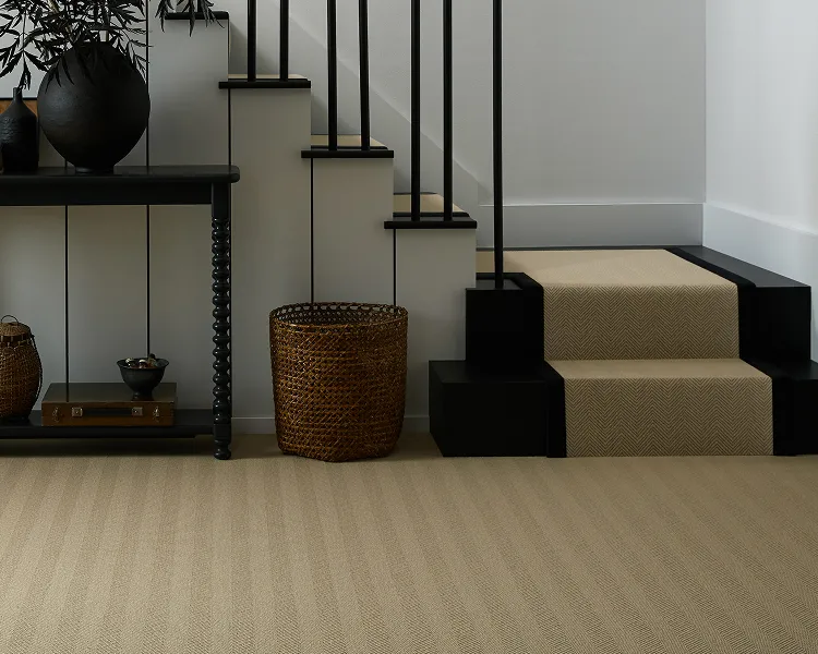 Entryway with beige carpeted stairs and black railings on the right, white walls. A black console table on the left holds wicker baskets, a small bowl and a tall plant.