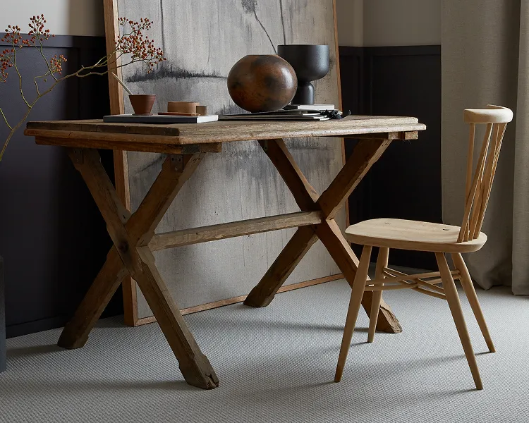 Rustic wooden trestle table with a round dark vase, small bowls and books, beside a light chair; an abstract panel rests behind in a cozy room.