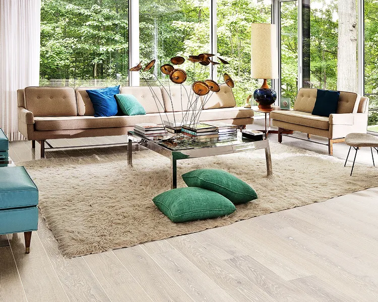 Bright modern living room with big windows, beige sofas, blue/green cushions, glass coffee table with a dried flower centrepiece, and a shag rug.