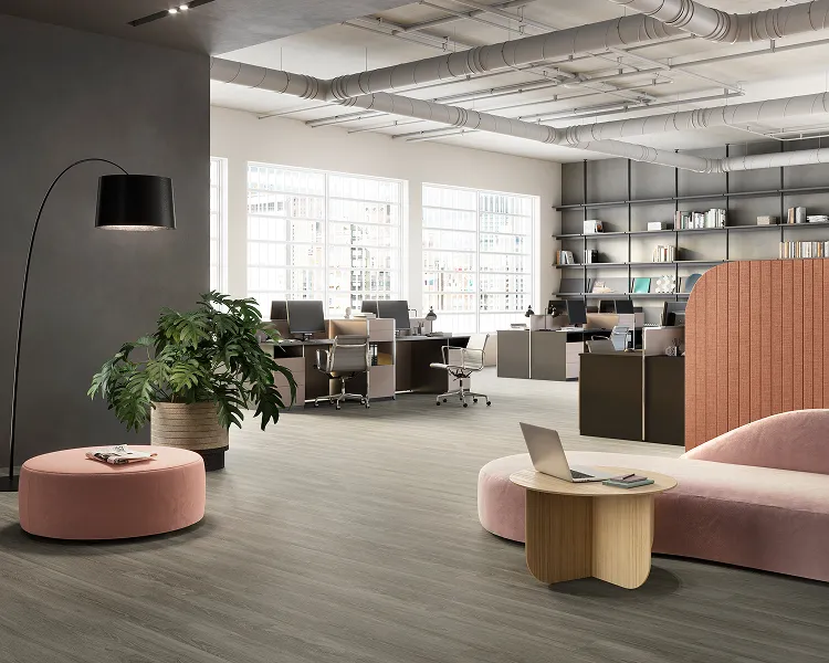 Open-plan office lounge with a pink curved sofa and ottoman, small wooden table with a laptop, a leafy plant, and desks in the background under exposed ceiling pipes.