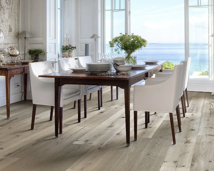 Bright dining room with a dark wooden table set with plates, glassware and a vase of flowers; white chairs surround it as large windows overlook the sea.
