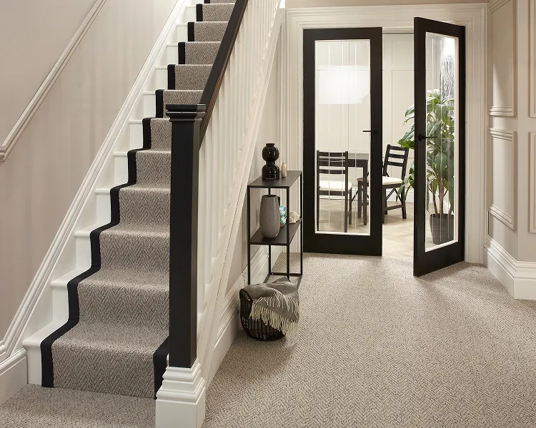 Hallway with a carpeted staircase on the left, black banister, and glass-panel doors opening to a dining area with a plant.