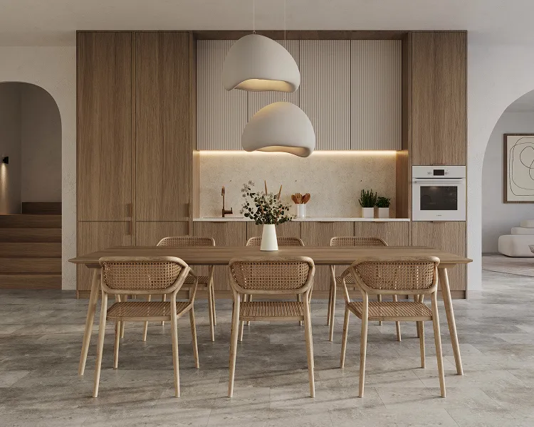 Open-plan kitchen and dining area with a long wooden table and six woven-back chairs, warm wood cabinetry, a built-in oven, and two sculptural white pendant lights.