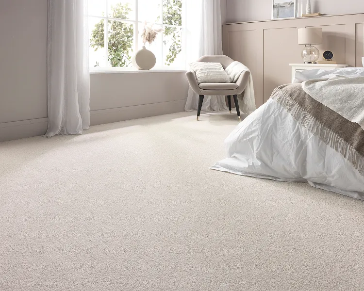 Bright bedroom with cream carpet, white bedding and a beige throw. A chair by a sunlit window, framed by sheer curtains and a vase on the sill.