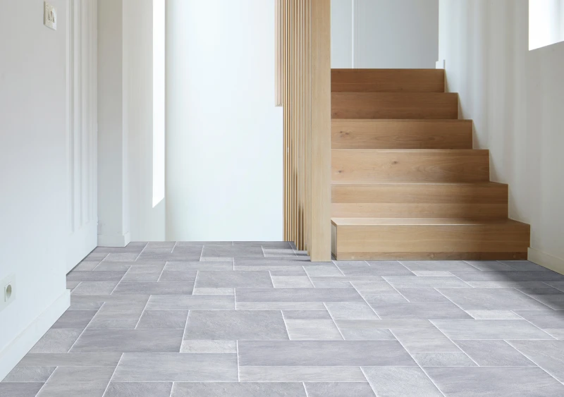 Bright modern interior with grey rectangular floor tiles, white walls, and a light wooden staircase on the right, separated by a vertical slatted divider.