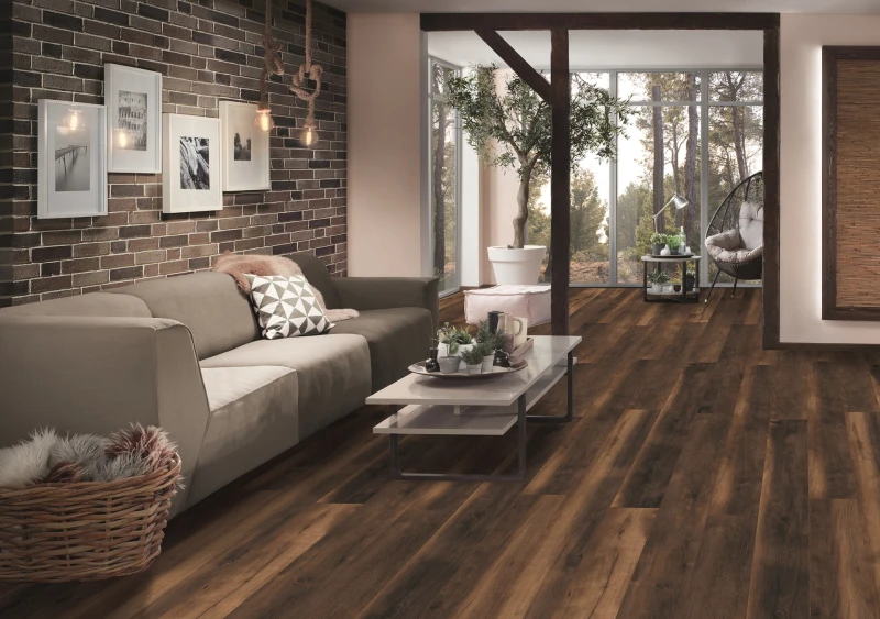 Modern living room with a brick wall, grey sofa with geometric cushion, rope pendant lights, a wicker basket, white coffee table, and large glass doors opening to a garden.