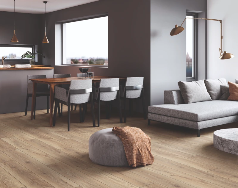 Open-plan living and dining area with a wooden table and grey chairs, a grey sofa, warm wood floors, brass floor lamp, a pouf with an orange throw, and large windows.