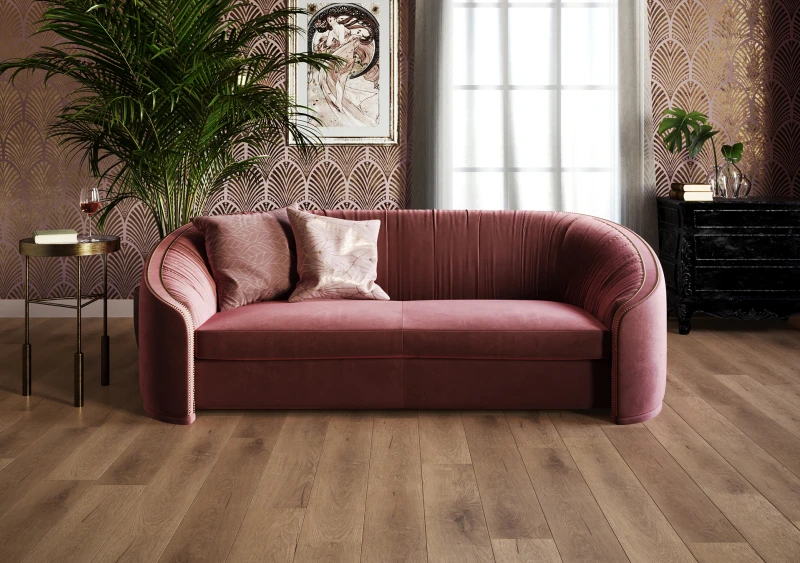 Pink velvet curved sofa with two cushions in a retro-styled room; tall plant, patterned wallpaper, side table with a wine glass, framed art, and a black dresser by a window.