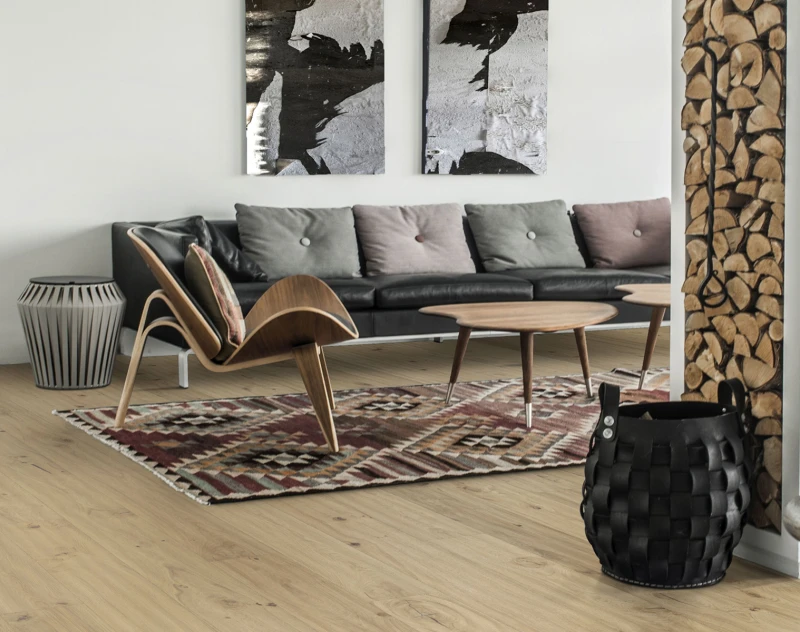 Modern living room with a black sofa, pastel cushions, curved wooden lounge chair, round coffee tables, rug, stacked firewood, and a woven basket.