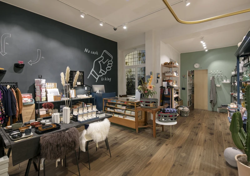 Interior of a modern boutique with a dark chalkboard wall displaying No cash is king, wooden floors, clothing rails, display tables, shelves and cosy seating.