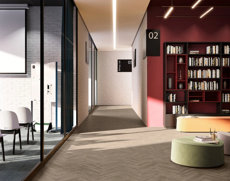 Bright modern library corridor with white brick walls, glass partition left, burgundy bookshelf wall right, and green and orange seating up front.