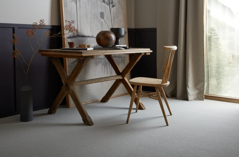 Rustic wooden desk with cross-leg frame and a round sculpture; a wooden chair beside a tall vase with berries, next to a window with beige curtains.