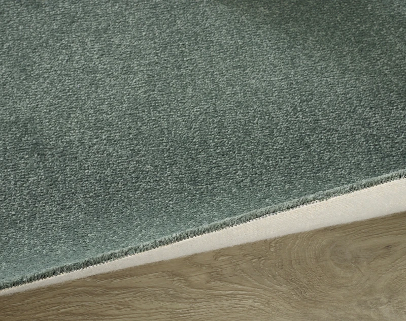 Close-up of a green textured carpet edge transitioning to a light wood floor, showing the beige underside and a diagonal seam.